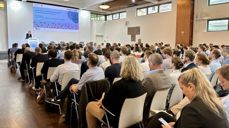 Christina Decker, Abteilungsleiterin Digital- und Innovationspolitik im BMWK bei ihrer Begrüßung Ein Sall voller Menschen auf Stühlen. Vorn eine Leinwand auf die eine Folie projeziert wird. Christina Decker, Abteilungsleiterin Digital- und Innovationspolitik im BMWK, spricht.