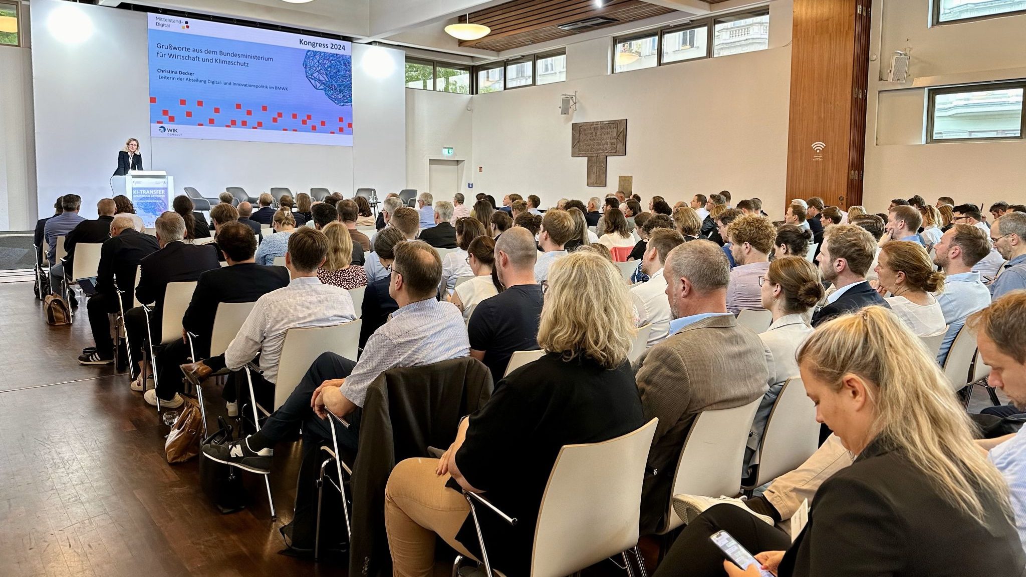 Ein Sall voller Menschen auf Stühlen. Vorn eine Leinwand auf die eine Folie projeziert wird. Christina Decker, Abteilungsleiterin Digital- und Innovationspolitik im BMWK, spricht.