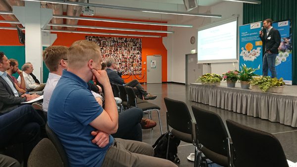 Ein Mann auf einer Bühne mit einem Mikrofon in der Hand spricht zu Teilnehmern