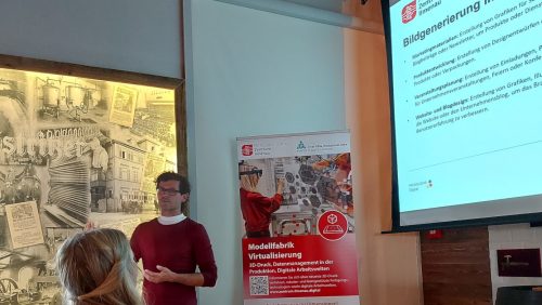 Karsten Jahn vom Mittelstand-Digital Zentrum Ilmenau bei seinem Vortrag über generative KI im Büromanagement. Rechts neben ihm befindet sich ein Aufsteller der Modellfabrig Virtualisierung. Am rechten Rand des Bildes ist eine Folie seines Vortrages zu sehen.