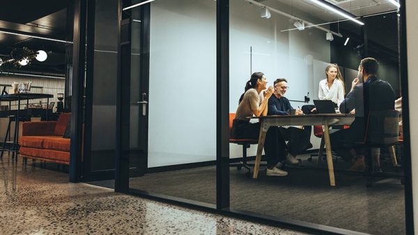 Menschen arbeiten an einem Tisch zusammen. Dabei sitzen sie in einem abgetrennten Bereich aus Glas.