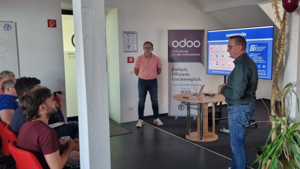 Vortrag Referenten, ein Bildschirm und sitzend im Vordergrund Gäste