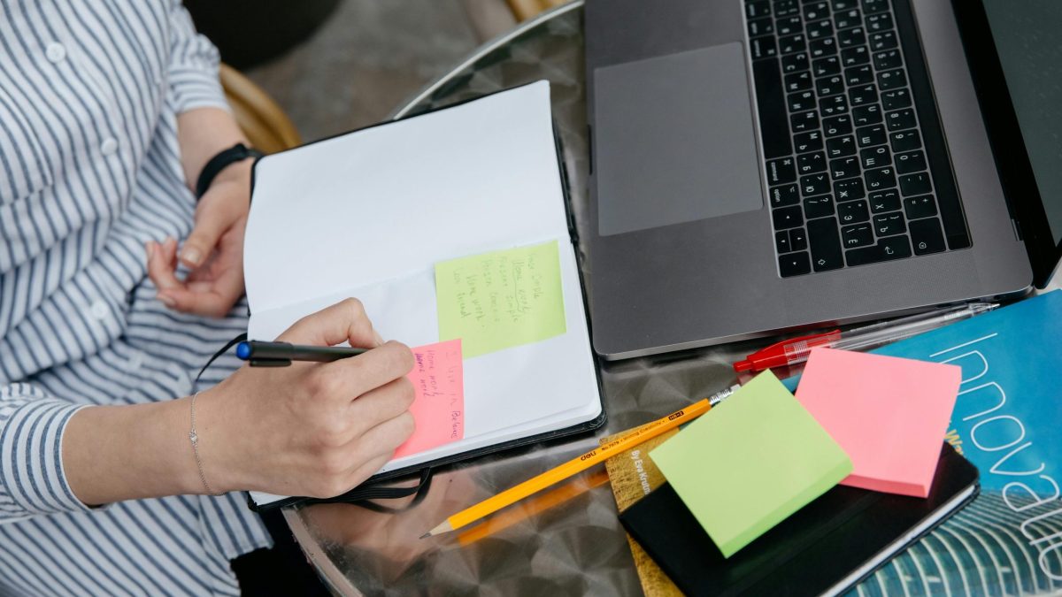 Schreibtisch Eine Person sitzt an einem Schreibtisch. Darauf ein Laptop und Notizzettel. Die Person macht handschriftliche Noitzen