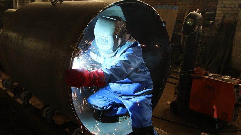Person im Schutzanzug sitzt mit Schweißermaske in einer Röhre und schweißt.