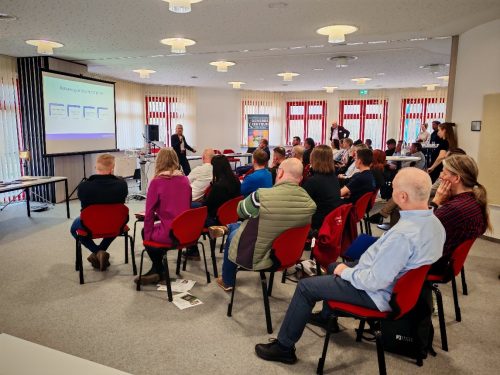 Viele Menschen sitzen auf Stühlen und schauen auf eine Beamerleinwand, die eine Präsentation zeigt