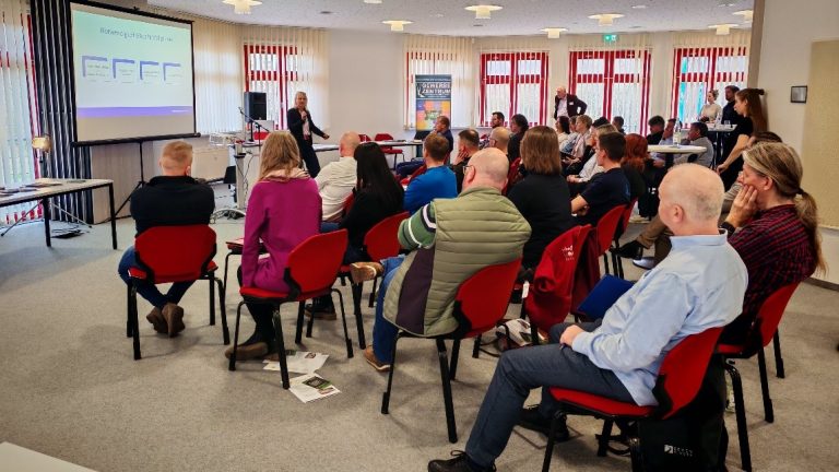 Viele Menschen sitzen auf Stühlen und schauen auf eine Beamerleinwand, die eine Präsentation zeigt