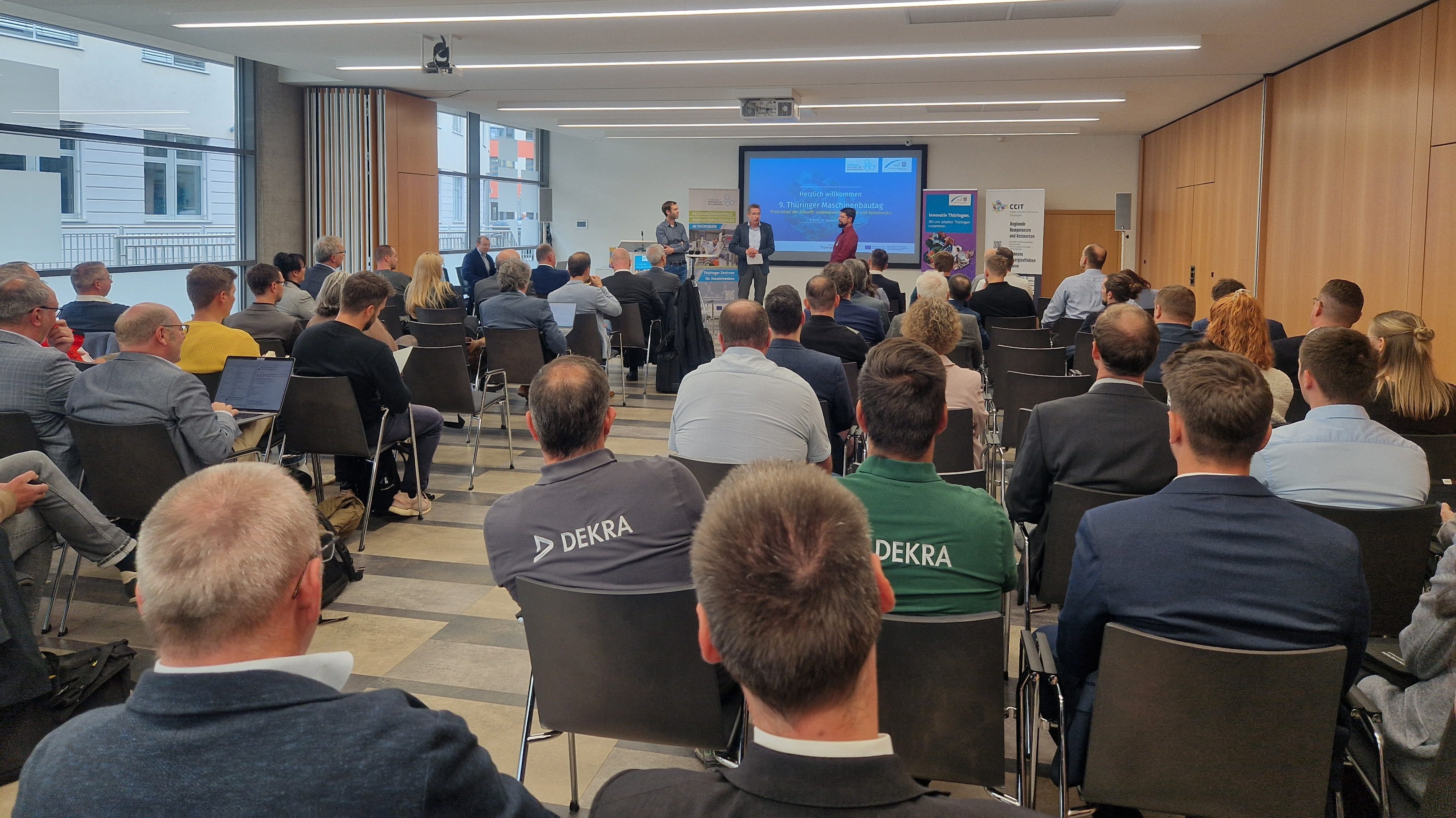 Viele Personen sitzen in einem Saal und schauen auf einen Redner