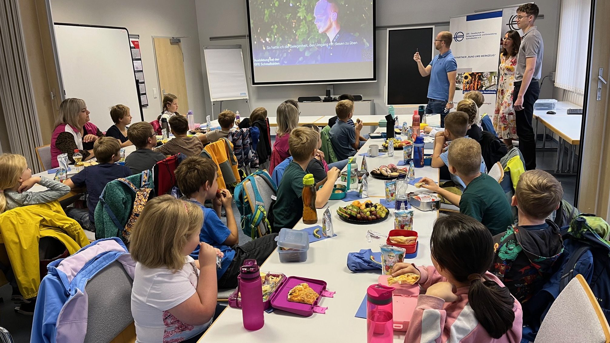 Gemeinsam lernen Eine Gruppe von Grundschulkindern sitzt an langen Tischen in einem Klassenraum und schaut nach vorne zu drei Erwachsenen, die vor einer Leinwand präsentieren. Auf den Tischen stehen Brotdosen, Trinkflaschen und Obstteller. Die Kinder wirken aufmerksam und essen gleichzeitig ihre Pausenmahlzeit