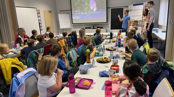 Gemeinsam lernen Eine Gruppe von Grundschulkindern sitzt an langen Tischen in einem Klassenraum und schaut nach vorne zu drei Erwachsenen, die vor einer Leinwand präsentieren. Auf den Tischen stehen Brotdosen, Trinkflaschen und Obstteller. Die Kinder wirken aufmerksam und essen gleichzeitig ihre Pausenmahlzeit