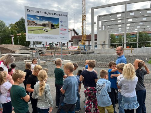 Eine Gruppe von Kindern steht vor einer Baustelle, im Hintergrund ein Baustellenplakat mit dem Titel Zentrum für digitale Produktion.