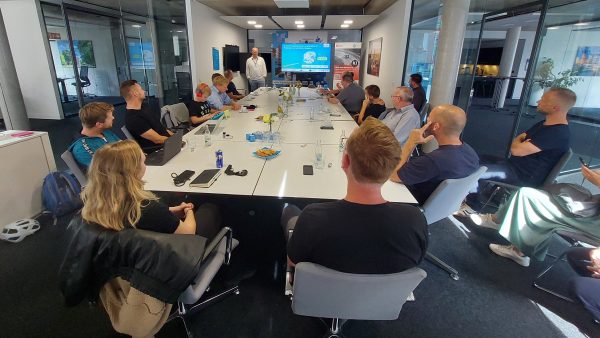 KI-Jetzt! Menschen sitzen um einen Tisch herum und schauen auf einen Referenten vor einem Beamer-Bild