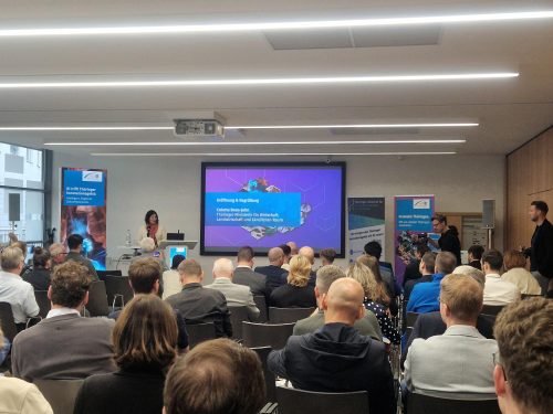 Im Konferenzraum des 7. Thüringer KI-Forums hält eine Rednerin am Pult den Eröffnungsvortrag vor einem Publikum. Auf der Leinwand steht „Eröffnung & Begrüßung – Colette Böhm-Holm, Thüringer Ministerium für Wirtschaft, Landwirtschaft und ländlichen Raum“. Rund 40 Teilnehmende sitzen im Saal, umgeben von Bannern zum Thema Innovation und Künstliche Intelligenz.