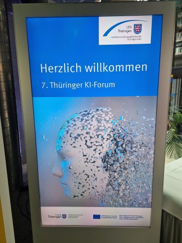 Digitale Anzeigetafel mit der Aufschrift „Herzlich willkommen – 7. Thüringer KI-Forum“ und dem Logo der Landesentwicklungsgesellschaft Thüringen (LEG). Im unteren Bereich ist eine stilisierte, aus Pixeln aufgelöste menschliche Kopf-Silhouette zu sehen.