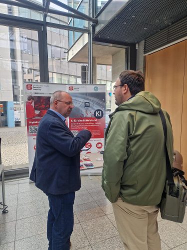 Zwei Männer führen ein Gespräch am Informationsstand des Mittelstand-Digital Zentrums Ilmenau. Im Hintergrund ist ein Roll-up mit Informationen zu KI-Anwendungen für den Mittelstand zu sehen.
