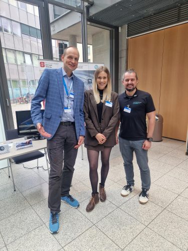 Drei Personen stehen lächelnd an einem Messestand des Mittelstand-Digital Zentrums Ilmenau. Hinter ihnen sind Banner zum Thema Künstliche Intelligenz und Digitalisierung zu sehen.