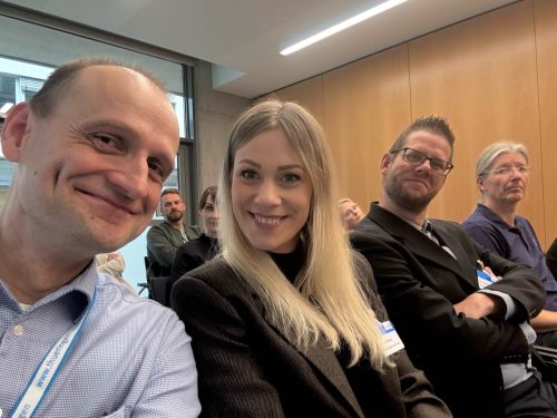 Personen sitzen in einer Konferenzreihe und lächeln in die Kamera. Im Hintergrund sind weitere Teilnehmende zu sehen, die dem Veranstaltungsgeschehen folgen.