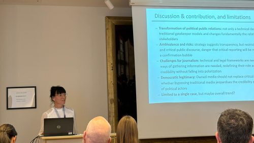 Dr. Ninette Florschütz hält einen wissenschaftlichen Vortrag vor Publikum. Auf der Leinwand ist eine Präsentationsfolie mit dem Titel „Discussion & contribution, and limitations“ zu sehen, die Punkte zu politischer Öffentlichkeitsarbeit, Journalismus und demokratischer Legitimation enthält.