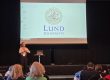 Eine Person steht auf einer Bühne und spricht vor einem Publikum. Auf der Leinwand hinter ihr ist das Logo der „Lund University“ mit dem Universitätswappen zu sehen. Im Vordergrund sitzen mehrere Zuhörerinnen und Zuhörer.