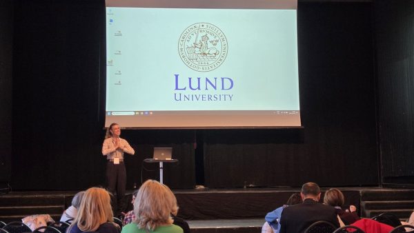 Vortrag Lund University Eine Person steht auf einer Bühne und spricht vor einem Publikum. Auf der Leinwand hinter ihr ist das Logo der „Lund University“ mit dem Universitätswappen zu sehen. Im Vordergrund sitzen mehrere Zuhörerinnen und Zuhörer.