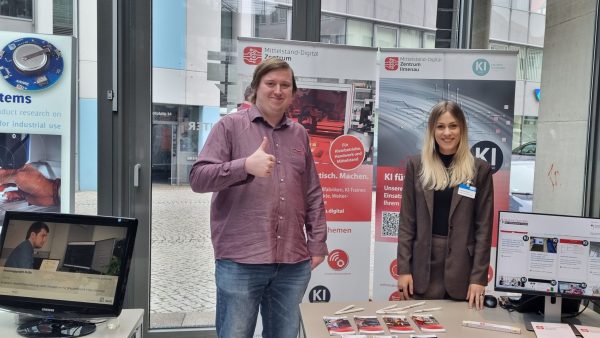 Thüringer KI-Forum – Vernetzung Zwei Personen stehen an einem Infostand des Mittelstand-Digital Zentrums Ilmenau. Hinter ihnen sind Roll-up-Banner mit Informationen zu Künstlicher Intelligenz für kleine und mittlere Unternehmen zu sehen. Auf dem Tisch liegen Flyer und Messutensilien, daneben steht ein Monitor mit einer Präsentation.
