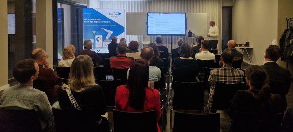 Informationsveranstaltung zum digitalen Wandel in Thüringen Eine Gruppe von Teilnehmenden sitzt in einem modernen Seminarraum und hört einem Vortragenden zu, der vorn neben einem Bildschirm steht. Auf dem Bildschirm ist die Agenda der Veranstaltung zu sehen. Im Hintergrund steht ein Roll-up mit der Aufschrift „Wir gestalten mit euch den digitalen Wandel. Für kleine und mittlere Unternehmen in Thüringen“ von ThEx Thüringen.