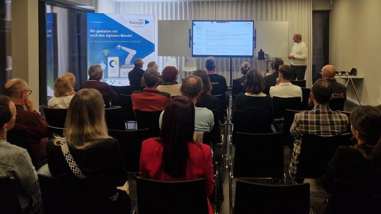 Informationsveranstaltung zum digitalen Wandel in Thüringen Eine Gruppe von Teilnehmenden sitzt in einem modernen Seminarraum und hört einem Vortragenden zu, der vorn neben einem Bildschirm steht. Auf dem Bildschirm ist die Agenda der Veranstaltung zu sehen. Im Hintergrund steht ein Roll-up mit der Aufschrift „Wir gestalten mit euch den digitalen Wandel. Für kleine und mittlere Unternehmen in Thüringen“ von ThEx Thüringen.