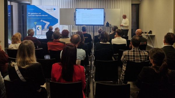 Informationsveranstaltung zum digitalen Wandel in Thüringen Eine Gruppe von Teilnehmenden sitzt in einem modernen Seminarraum und hört einem Vortragenden zu, der vorn neben einem Bildschirm steht. Auf dem Bildschirm ist die Agenda der Veranstaltung zu sehen. Im Hintergrund steht ein Roll-up mit der Aufschrift „Wir gestalten mit euch den digitalen Wandel. Für kleine und mittlere Unternehmen in Thüringen“ von ThEx Thüringen.