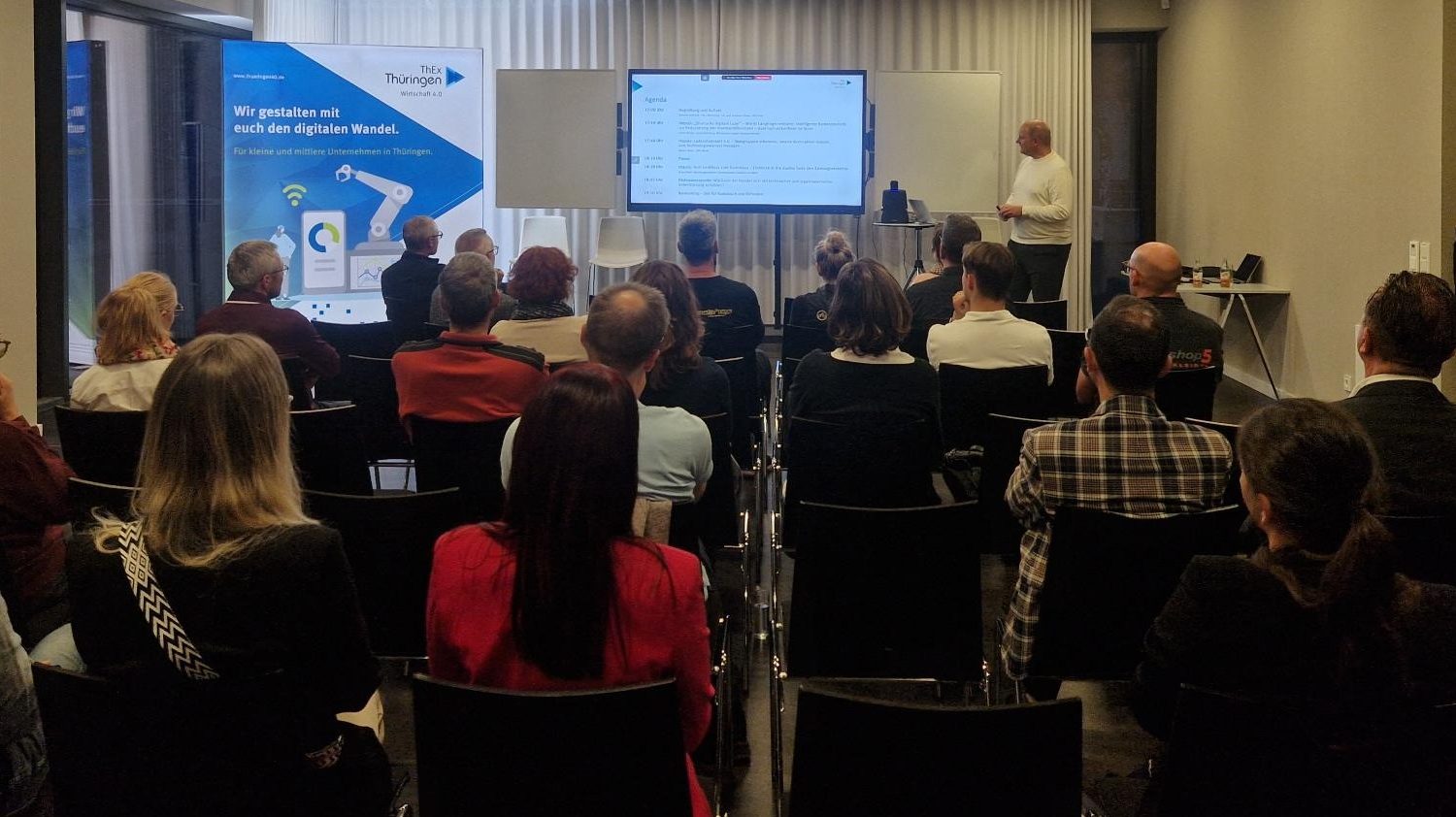 Eine Gruppe von Teilnehmenden sitzt in einem modernen Seminarraum und hört einem Vortragenden zu, der vorn neben einem Bildschirm steht. Auf dem Bildschirm ist die Agenda der Veranstaltung zu sehen. Im Hintergrund steht ein Roll-up mit der Aufschrift „Wir gestalten mit euch den digitalen Wandel. Für kleine und mittlere Unternehmen in Thüringen“ von ThEx Thüringen.