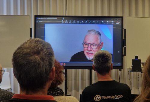 Das Bild zeigt eine Gruppe von Personen, die in einem Seminar- oder Konferenzraum sitzen und gemeinsam auf einen Bildschirm schauen. Auf dem Bildschirm ist ein Mann mit Brille zu sehen, der offenbar per Videokonferenz zugeschaltet ist und spricht. Der Raum ist modern eingerichtet, mit hellen Vorhängen und einer schlichten Beleuchtung.