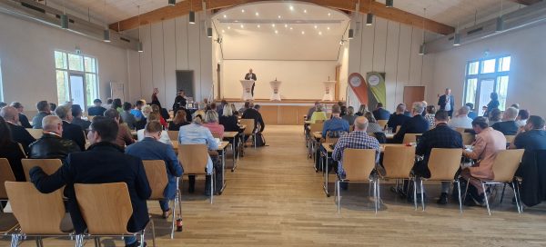 Regionalkonferenz des Thüringer Bogens In einem großen Veranstaltungssaal mit Holzdecke und heller Beleuchtung sitzen zahlreiche Teilnehmende an Tischen und hören einem Redner zu, der auf einer Bühne hinter einem Stehpult spricht. Neben ihm stehen weitere Pulte und zwei Roll-Ups mit Logos