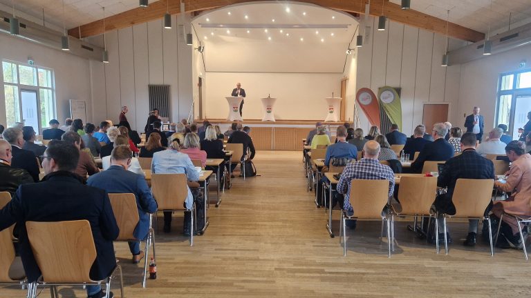 Regionalkonferenz des Thüringer Bogens In einem großen Veranstaltungssaal mit Holzdecke und heller Beleuchtung sitzen zahlreiche Teilnehmende an Tischen und hören einem Redner zu, der auf einer Bühne hinter einem Stehpult spricht. Neben ihm stehen weitere Pulte und zwei Roll-Ups mit Logos