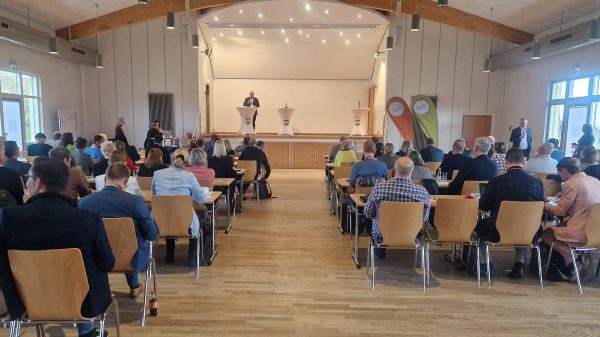 Regionalkonferenz des Thüringer Bogens In einem großen Veranstaltungssaal mit Holzdecke und heller Beleuchtung sitzen zahlreiche Teilnehmende an Tischen und hören einem Redner zu, der auf einer Bühne hinter einem Stehpult spricht. Neben ihm stehen weitere Pulte und zwei Roll-Ups mit Logos