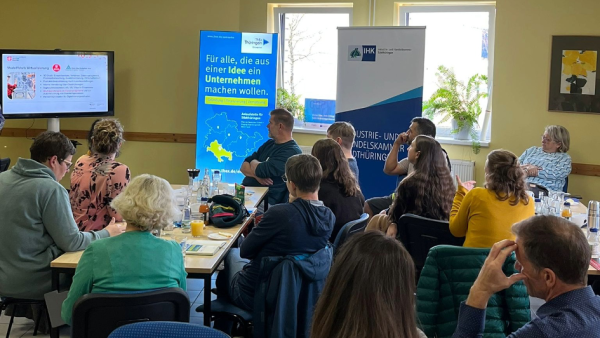 Smartes Büromanagement Zella Mehlis Das Bild zeigt eine Gruppe von Menschen, die in einem Seminarraum an einer Veranstaltung teilnehmen. Sie sitzen an Tischen, hören einer Präsentation zu und blicken auf einen Bildschirm, auf dem eine Folie zum Thema „Mobilitäts Virtualisierung“ angezeigt wird. Im Hintergrund stehen Roll-Ups mit Logos und dem Slogan: „Für alle, die aus einer Idee ein Unternehmen machen wollen.“