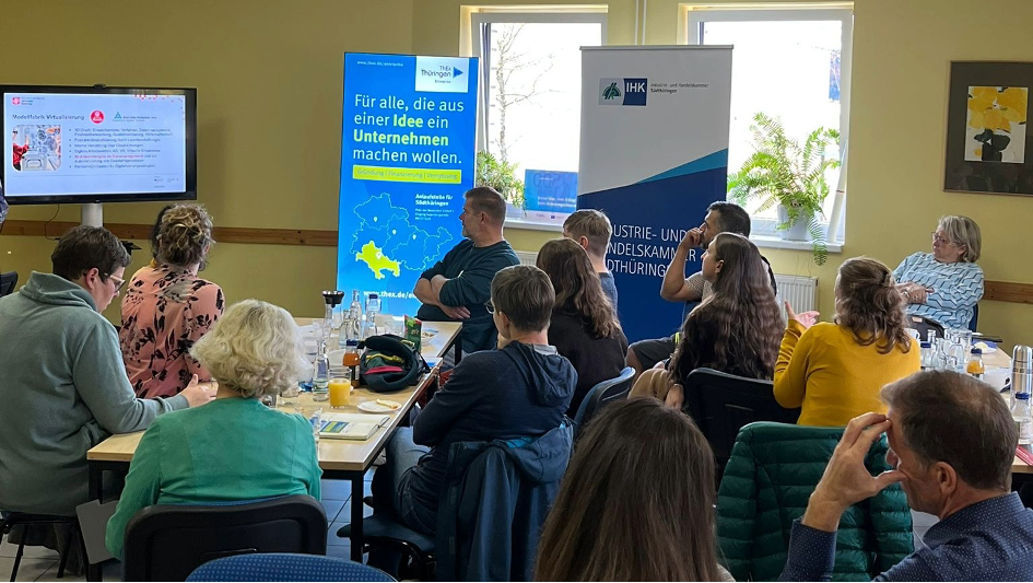 Das Bild zeigt eine Gruppe von Menschen, die in einem Seminarraum an einer Veranstaltung teilnehmen. Sie sitzen an Tischen, hören einer Präsentation zu und blicken auf einen Bildschirm, auf dem eine Folie zum Thema „Mobilitäts Virtualisierung“ angezeigt wird. Im Hintergrund stehen Roll-Ups mit Logos und dem Slogan: „Für alle, die aus einer Idee ein Unternehmen machen wollen.“