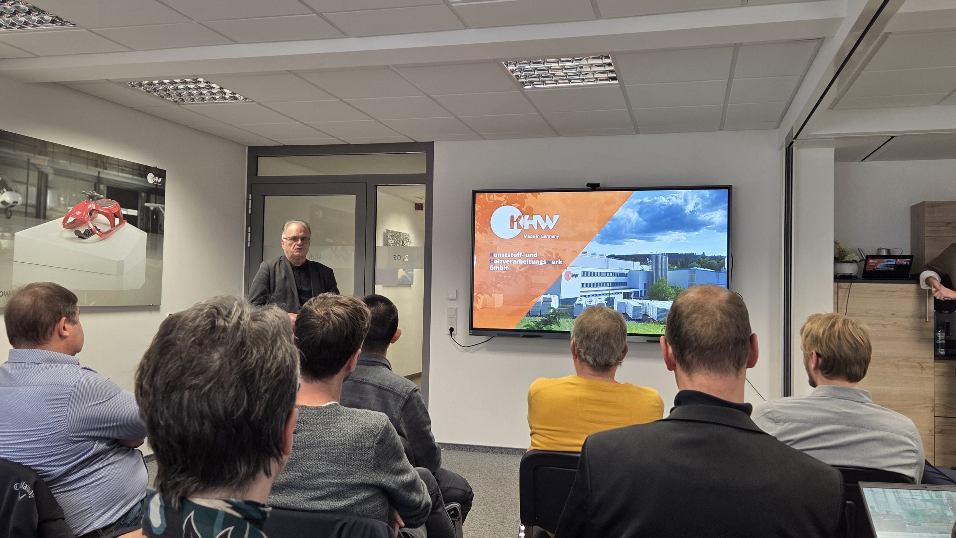 Eine Gruppe von Teilnehmenden sitzt in einem Seminarraum und hört einem Vortragenden zu, der vor einer Tür steht. Auf dem großen Bildschirm rechts wird eine Präsentationsfolie mit dem Logo und einem Gebäude des Unternehmens KHW angezeigt. Ein weiterer Mann lehnt rechts an einem Schrank und beobachtet die Szene.