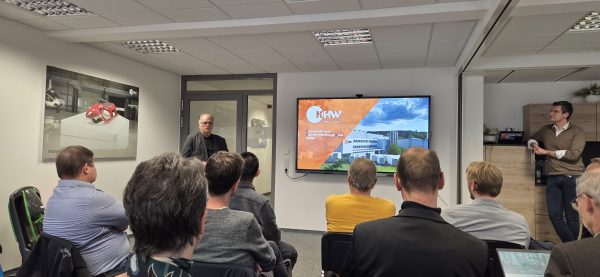 Eine Gruppe von Teilnehmenden sitzt in einem Seminarraum und hört einem Vortragenden zu, der vor einer Tür steht. Auf dem großen Bildschirm rechts wird eine Präsentationsfolie mit dem Logo und einem Gebäude des Unternehmens KHW angezeigt. Ein weiterer Mann lehnt rechts an einem Schrank und beobachtet die Szene.