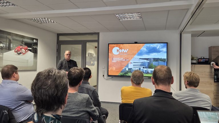 Eine Gruppe von Teilnehmenden sitzt in einem Seminarraum und hört einem Vortragenden zu, der vor einer Tür steht. Auf dem großen Bildschirm rechts wird eine Präsentationsfolie mit dem Logo und einem Gebäude des Unternehmens KHW angezeigt. Ein weiterer Mann lehnt rechts an einem Schrank und beobachtet die Szene.