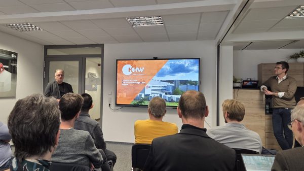 Unternehmenspräsentation Eine Gruppe von Teilnehmenden sitzt in einem Seminarraum und hört einem Vortragenden zu, der vor einer Tür steht. Auf dem großen Bildschirm rechts wird eine Präsentationsfolie mit dem Logo und einem Gebäude des Unternehmens KHW angezeigt. Ein weiterer Mann lehnt rechts an einem Schrank und beobachtet die Szene.