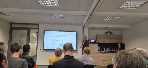 Eine Gruppe von Zuhörenden sitzt in einem Seminarraum und verfolgt einen Vortragenden, der vor einem großen Bildschirm steht und Forschungsthemen sowie ein Prozessdiagramm präsentiert. Rechts im Raum befindet sich eine kleine Kaffeestation mit Getränken.
