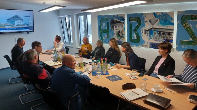 Eine Gruppe von etwa zwölf Personen sitzt in einem hellen Besprechungsraum an einem langen Konferenztisch. Auf dem Tisch stehen Wasserflaschen, Gläser, Kaffeetassen und Notizmaterial. Die Teilnehmenden hören einer Präsentation zu, die auf einem Bildschirm an der Wand gezeigt wird. An der Wand hängen große, farbige Bildtafeln mit abstrakten Motiven und Gebäuden. Tageslicht fällt durch mehrere Fenster in den Raum.