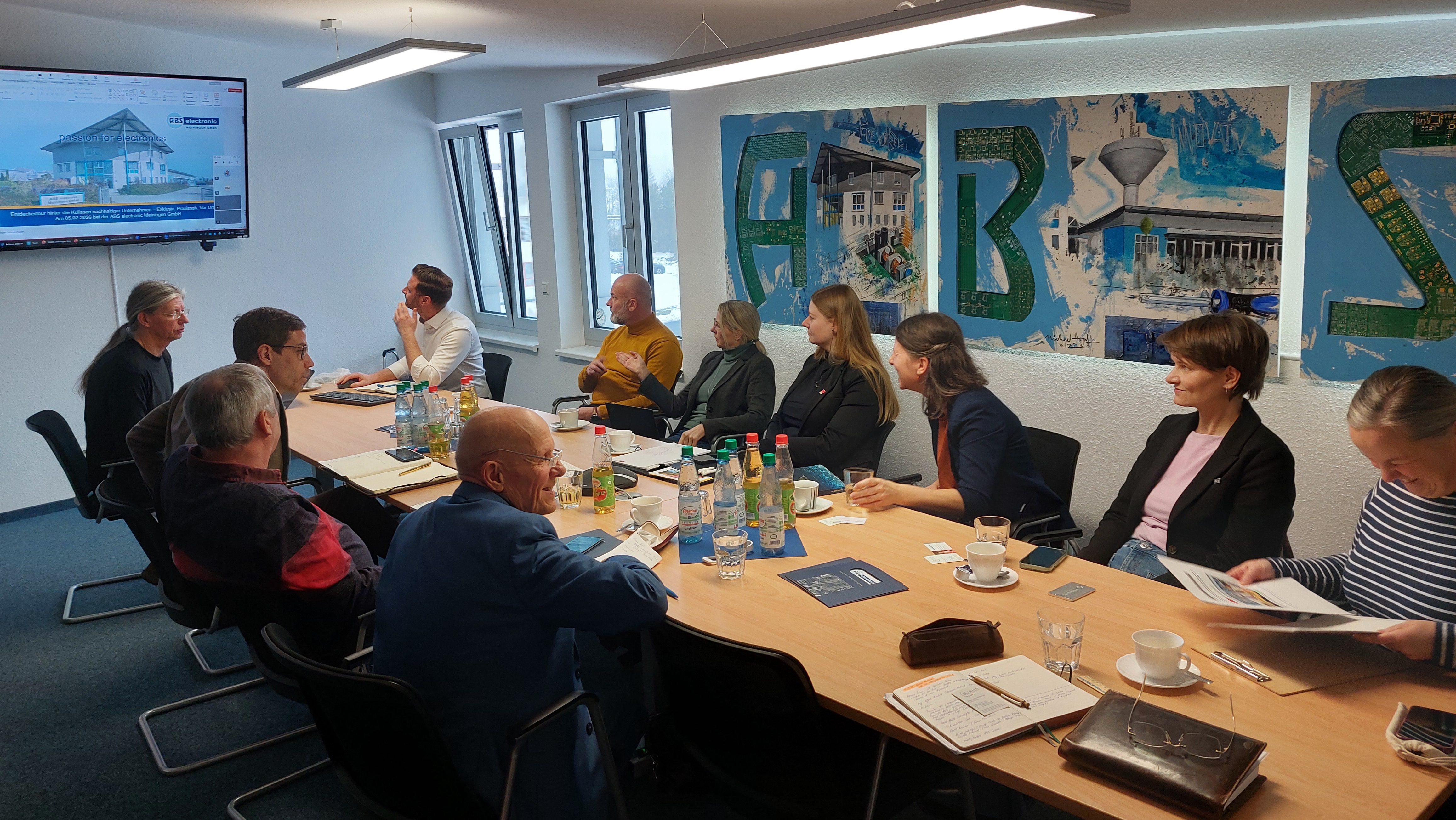 Eine Gruppe von etwa zwölf Personen sitzt in einem hellen Besprechungsraum an einem langen Konferenztisch. Auf dem Tisch stehen Wasserflaschen, Gläser, Kaffeetassen und Notizmaterial. Die Teilnehmenden hören einer Präsentation zu, die auf einem Bildschirm an der Wand gezeigt wird. An der Wand hängen große, farbige Bildtafeln mit abstrakten Motiven und Gebäuden. Tageslicht fällt durch mehrere Fenster in den Raum.
