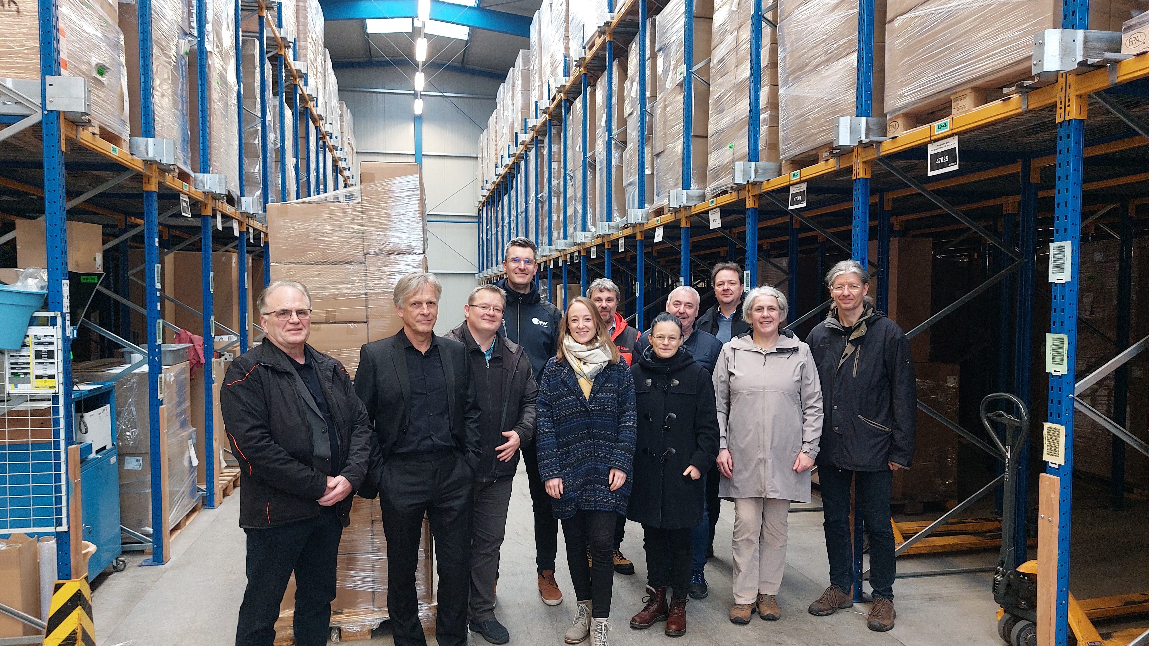 Gruppenfoto in der Produktion Eine Gruppe von zehn Personen steht in einer Lagerhalle zwischen hohen Regalen mit palettierten und in Folie verpackten Kartons. Die Regale reichen bis unter die Hallendecke und sind beidseitig angeordnet. Die Personen posieren in der Mitte des Gangs für ein Gruppenfoto. Im Hintergrund sind weitere gestapelte Waren sowie ein Hubwagen zu sehen.