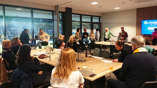 **Alt-Text:** Mehrere Personen sitzen und stehen in einem modern gestalteten Veranstaltungsraum um große Holztische herum. Eine Person spricht vor der Gruppe und gestikuliert, während andere zuhören oder Notizen machen. Im Hintergrund ist ein Bildschirm mit einer Präsentationsfolie zu sehen. Durch die großen Fenster erkennt man eine beleuchtete Sport- oder Veranstaltungshalle. Auf den Tischen stehen Getränke, Gläser und Unterlagen.