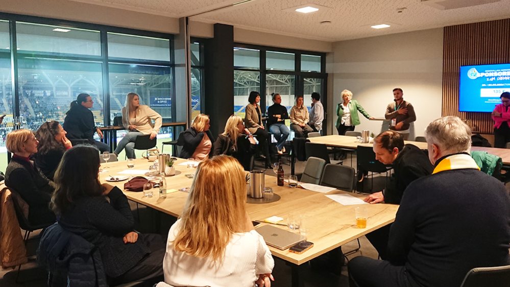 **Alt-Text:** Mehrere Personen sitzen und stehen in einem modern gestalteten Veranstaltungsraum um große Holztische herum. Eine Person spricht vor der Gruppe und gestikuliert, während andere zuhören oder Notizen machen. Im Hintergrund ist ein Bildschirm mit einer Präsentationsfolie zu sehen. Durch die großen Fenster erkennt man eine beleuchtete Sport- oder Veranstaltungshalle. Auf den Tischen stehen Getränke, Gläser und Unterlagen.