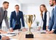 Ein goldener Pokal steht aufrecht und mittig auf einem hellen Konferenztisch in einem Büro. Im Hintergrund stehen mehrere Geschäftspersonen in Anzügen um den Tisch herum und beugen sich leicht nach vorn, als würden sie diskutieren. Der Raum ist hell ausgeleuchtet und wirkt wie ein professionelles Arbeitsumfeld.
