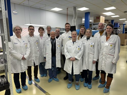 Eine Gruppe von 12 Personen steht nebeneinander in einer Produktions- oder Laborumgebung. Alle tragen weiße Laborkittel, Namensschilder und blaue Überziehschuhe. Im Hintergrund sind Regale, Maschinen und Kartons zu sehen, die auf einen technischen oder industriellen Arbeitsplatz hinweisen. Die Personen blicken in die Kamera und posieren für ein Gruppenfoto unter heller Deckenbeleuchtung.