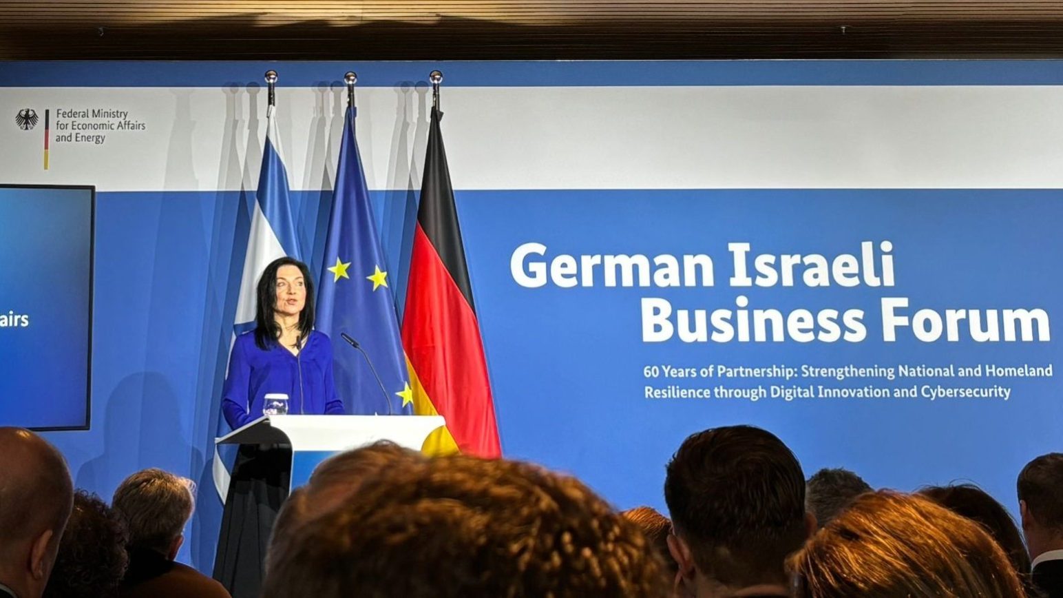 Katharina Reiche steht an einem Rednerpult und hält eine Ansprache vor Publikum. Hinter ihr sind die Flaggen Deutschlands, der Europäischen Union und Israels zu sehen. Auf einer großen blauen Wand im Hintergrund steht „German Israeli Business Forum“ mit einem Untertitel zur Partnerschaft und digitalen Innovation. Auf einem Bildschirm links neben der Rednerin wird ihr Name und ihre Funktion als Bundesministerin für Wirtschaft und Energie angezeigt. Im Vordergrund sind die Köpfe der Zuhörenden zu sehen.