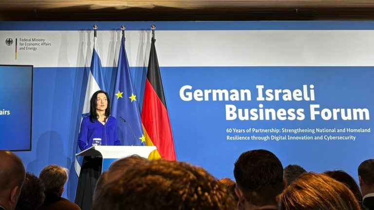 Katharina Reiche steht an einem Rednerpult und hält eine Ansprache vor Publikum. Hinter ihr sind die Flaggen Deutschlands, der Europäischen Union und Israels zu sehen. Auf einer großen blauen Wand im Hintergrund steht „German Israeli Business Forum“ mit einem Untertitel zur Partnerschaft und digitalen Innovation. Auf einem Bildschirm links neben der Rednerin wird ihr Name und ihre Funktion als Bundesministerin für Wirtschaft und Energie angezeigt. Im Vordergrund sind die Köpfe der Zuhörenden zu sehen.