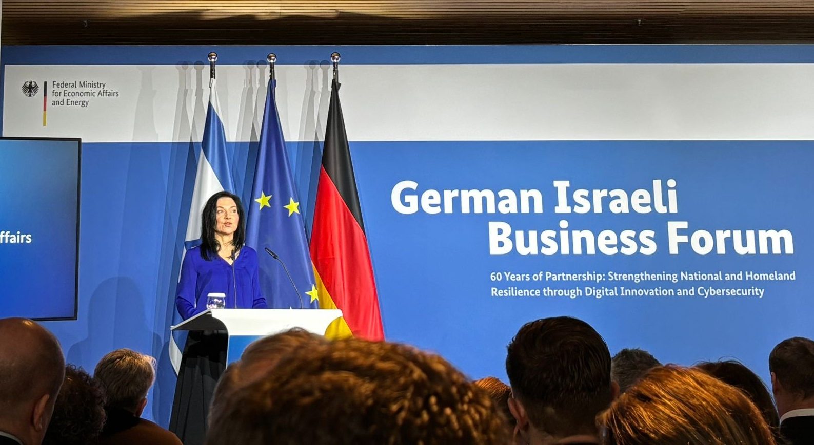Katharina Reiche steht an einem Rednerpult und hält eine Ansprache vor Publikum. Hinter ihr sind die Flaggen Deutschlands, der Europäischen Union und Israels zu sehen. Auf einer großen blauen Wand im Hintergrund steht „German Israeli Business Forum“ mit einem Untertitel zur Partnerschaft und digitalen Innovation. Auf einem Bildschirm links neben der Rednerin wird ihr Name und ihre Funktion als Bundesministerin für Wirtschaft und Energie angezeigt. Im Vordergrund sind die Köpfe der Zuhörenden zu sehen.
