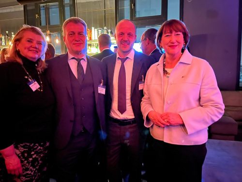 Vier Personen stehen nebeneinander bei einer Veranstaltung und blicken in die Kamera. Zwei Männer tragen Anzüge mit Krawatte und Namensschilder, die beiden Frauen sind ebenfalls formell gekleidet. Im Hintergrund ist eine Bar mit beleuchteten Flaschen und warmem, farbigem Licht zu sehen, das die Szene in Rot- und Blautöne taucht.